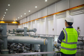 Worker in safety helmet and reflective vest inspects insulated gas switchgear substation indoors with large pipes and industrial equipment under bright lighting