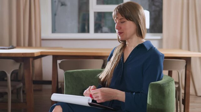 Wellness guide seated in green chair speaking intentionally to guest while holding pen and notebook on lap, warm modern therapy room with wooden table and muted curtains visible in background