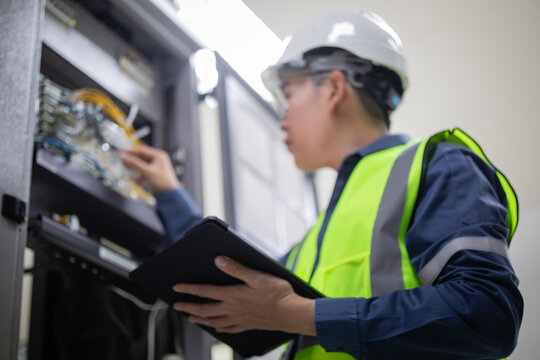 Network technician wearing safety helmet and reflective vest performs LAN patchpanel inspection using tablet in server room with focused attention and professional care