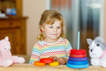 Cute little toddler girl playing alone with colorful wooden rainbow pyramid and toys at home or nursery. Happy healthy child having fun in kindergarten or preschool daycare