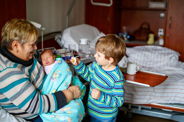 Happy grandmother holding newborn baby in hospital with proud big brother nearby. Real life, real...