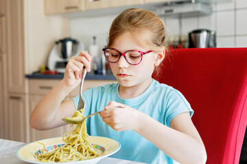 Little school girl eating spaghetti pasta with green pesto at home. Cute happy child with eyeglasses having healthy lunch.