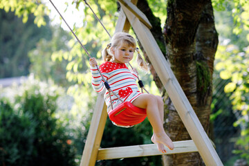 Happy beautiful little toddler girl having fun on swing in domestic garden. Cute healthy child swinging under blooming trees on sunny spring day. Baby laughing and crying