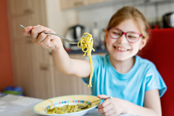 Little school girl eating spaghetti pasta with green pesto at home. Cute happy child with eyeglasses having healthy lunch.