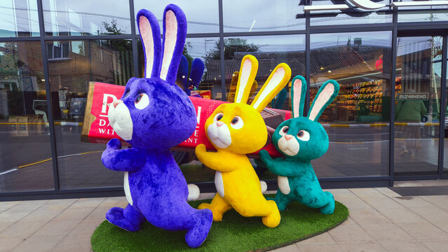 Colorful plush rabbits carrying a giant chocolate bar in a busy city environment during the day, Roshen sweets shop, Bucha, Ukraine 29.06.2025