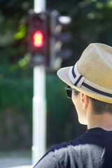 Moment from a journey to a city: a tourist in a hat waits at a pedestrian crossing with a red light in the background. Concepts of attentiveness and everyday charm of discovering a new destination