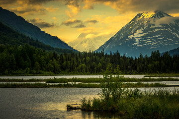The hidden sun is rising in the distance between two mountains coloring the sky gold with a lake in the foreground in Alaska.