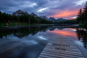 Photo of tranquil mountain lake at sunrise, fiery sky reflected on still water, jagged peaks, wooden dock with �Serenity Now� carving, evergreen silhouettes.