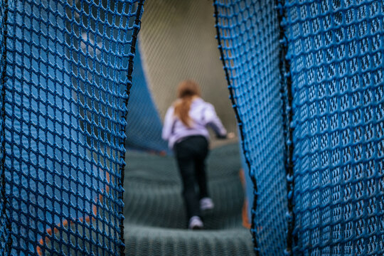 A young girl in a lavender hoodie climbs a soft net tunnel structure surrounded by blue mesh in an outdoor adventure park setting.