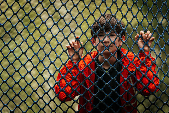 A young boy in a red jacket stands behind a blue rope safety net on an outdoor rope course, holding the net with both hands and looking through the openings.