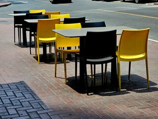 Side-by-side black and yellow chairs with tables on a brick sidewalk, bathed in sunlight. Ideal for outdoor dining or urban life concepts.
