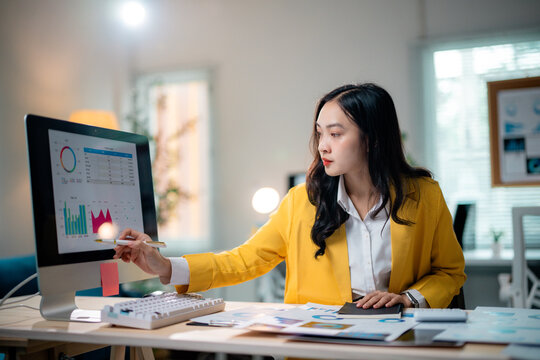 Asian businesswoman pointing at computer screen analyzing financial charts, working with documents in modern office
