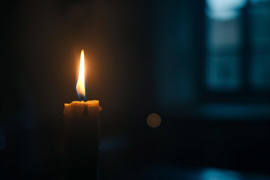A lit candle stands out against a darkened room. The flame emits a warm, soft light, contrasting with the blurry, dark background that features what seems to be a window.