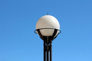 A contemporary white globe streetlamp is mounted on a black metal frame, standing tall and isolated...