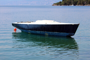 An old fiberglass boat with a faded blue hull floats quietly on still water, tethered to a buoy,...