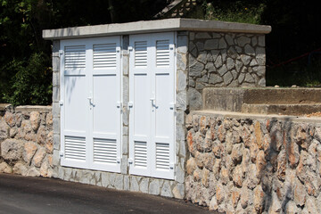 White metal utility cabinet with double louvered doors is installed in a stone wall, blending...