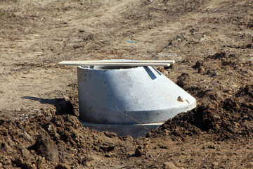 A concrete manhole base sits partially embedded in dry soil, topped with a wooden board, ready for...