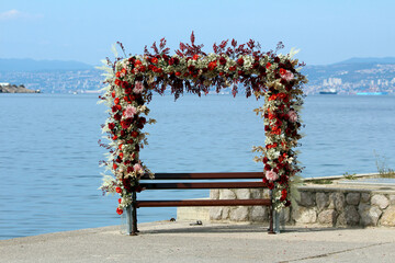 Wooden bench by the sea framed with arch of red rose rosa, white chrysanthemum chrysanthemum, and...