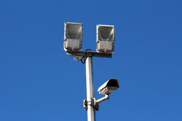 Two metal floodlights and a surveillance camera are fixed atop a tall pole, silhouetted against a...