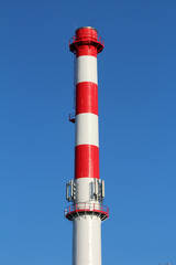 High industrial chimney with bold red and white stripes rises against a clear blue sky, equipped with antennas and safety railings for modern industry