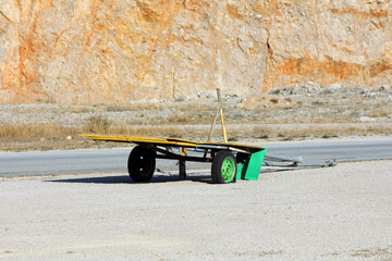 Yellow flatbed trailer with a green wheel and wooden frame sits on gravel beside a steep orange...