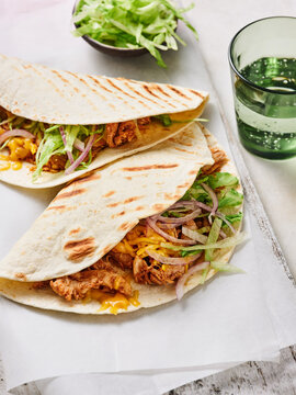 Two pulled pork burritos are presented on a light-colored platter. Coleslaw and fresh lettuce accompany the dish, along with a green drink. The setting is casual and inviting