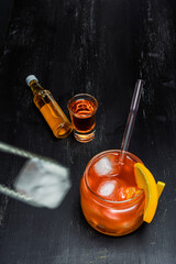 Jar of orange cocktail with ice cubes, garnished with orange slices and a glass straw, placed on a dark wooden surface. Nearby are a small bottle and shot glass filled with amber liquid, with ice tong