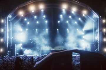 Nighttime concert scene with bright stage lighting, fog effects, and a large crowd in front of a DJ...