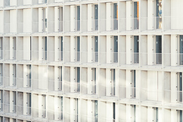 Side view of a modern apartment or hotel building with minimalist white balconies, glass doors, and repeating geometric architecture, illuminated by soft natural daylight and casting subtle shadows