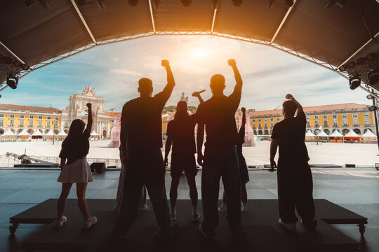 Backlit view of diverse performers silhouettes on stage raising fists in unity during Lisbon Pride event, celebrating LGBTQ+ rights and equality with iconic Arco da Rua Augusta in the background