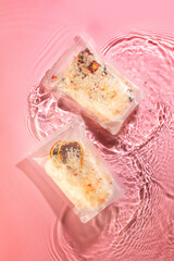 wo sealed packets of colorful food ingredients float gently in a rippling water pond on a soft pink background, creating a vibrant contrast. The ingredients within the packaging seem to be a blend of 