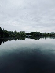 Grey cloudy sky reflection on the lake surface, silhouettes of the trees, lake surface as a mirror,...