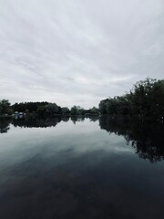 Grey cloudy sky reflection on the lake surface, silhouettes of the trees, lake surface as a mirror,...