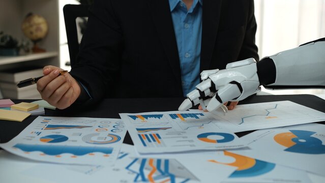 A businessman collaborates with a robotic arm to analyze financial reports, demonstrating the fusion of technology and human expertise in modern business environments.Noogenesis