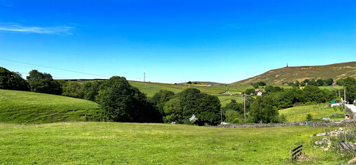 Rolling green hills stretch across the landscape under a clear blue sky. A few scattered trees and distant structures add to the serene countryside view in, Lumbutts, Todmorden, UK