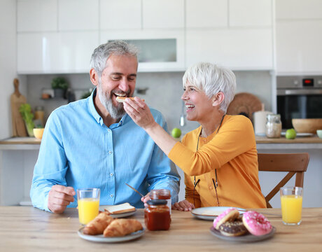 Portrait of an elderly senior couple having breakfast sharing a donut dessert at home. Happy healthy affectionate senior couple eating and sitting at kitchen table having fun enjoying morning meal tog