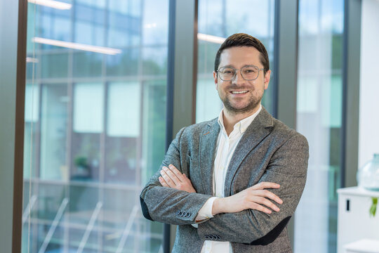 Portrait of a smiling young businessman standing in the office by the window in a suit and glasses, arms crossed on his chest and looking confidently at the camera