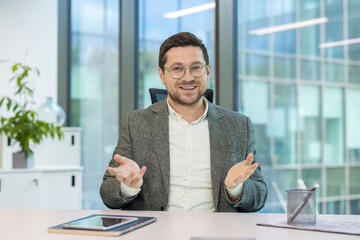 Fototapeta premium Smiling young male businessman sitting in the office at the table and holding an online conference via video call, gesturing with his hands at the camera