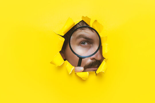 A young man with a mustache and beard looks through a magnifying glass (loupe) through a torn hole in a yellow paper. The concept of search, investigation, control,tax inspection and private detective
