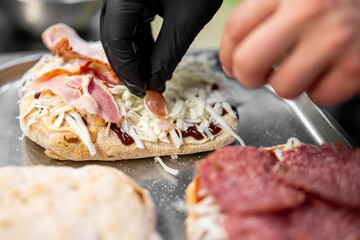 Close-up of a gloved hand preparing a sandwich with bacon, cheese, and sauce on a tray, showcasing fresh ingredients and street food preparation in a kitchen setting.
