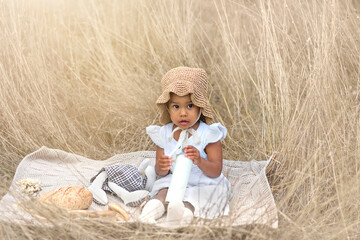 At a picnic, a small dark-skinned girl holds a bottle of milk and sits on a blanket with toys in a grain field