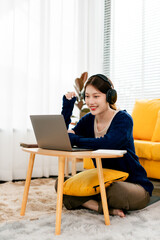 Beautiful Asian female student studying online at home. Young woman wearing earphones listening to music online.