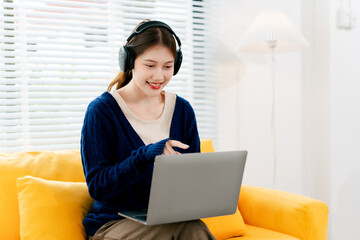 Naklejka premium Beautiful Asian female student studying online at home. Young woman wearing earphones listening to music online.