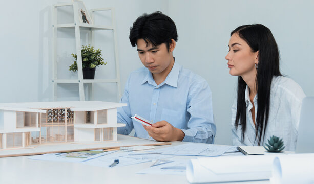 Professional male asian architect using ruler to measure house model length while young beautiful caucasian colleague using laptop to analyzed data on meeting table with house model. Immaculate.