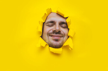 Laughing young man with closed eyes looking out through a torn hole on yellow paper. Concept of...