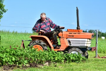 Senior farmer on compact orange tractor cultivating crops in lush green field. Summer agricultural...