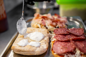 Close-up of sandwich preparation with toppings like salami, bacon, cheese, and sauce being added...