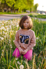 Child contemplating spent dandelion stem in field. Post-bloom moment, dark blonde hair. Pink pants, reflective childhood expression