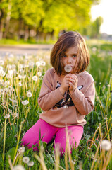 Child preparing to blow dandelion seeds, intense focus. Dark blonde hair glowing, pink leggings. Summer backlight, magical anticipation