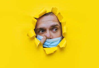 A man in a blue medical mask looks in surprise into a broken hole in yellow paper and picks his...